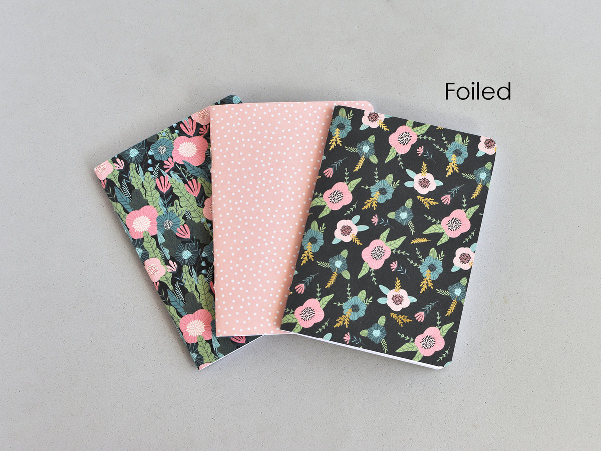 Three small notebooks or journal covers fanned out on a light gray background. The center cover is light pink with tiny white dots, and the two side covers are black with floral prints in pink, blue, green, and yellow.