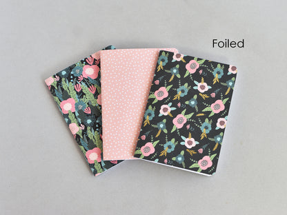 Three small notebooks or journal covers fanned out on a light gray background. The center cover is light pink with tiny white dots, and the two side covers are black with floral prints in pink, blue, green, and yellow.