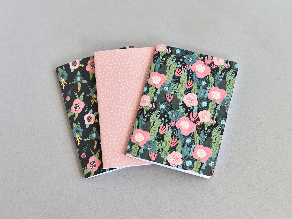 Three small notebooks or journals fanned out on a light gray background. The left notebook has a black background with pink and green floral pattern, the middle notebook is light pink with small white polka dots, and the right notebook has a dark background with pink and green floral pattern.