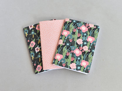 Three small notebooks or journals fanned out on a light gray background. The left notebook has a black background with pink and green floral pattern, the middle notebook is light pink with small white polka dots, and the right notebook has a dark background with pink and green floral pattern.