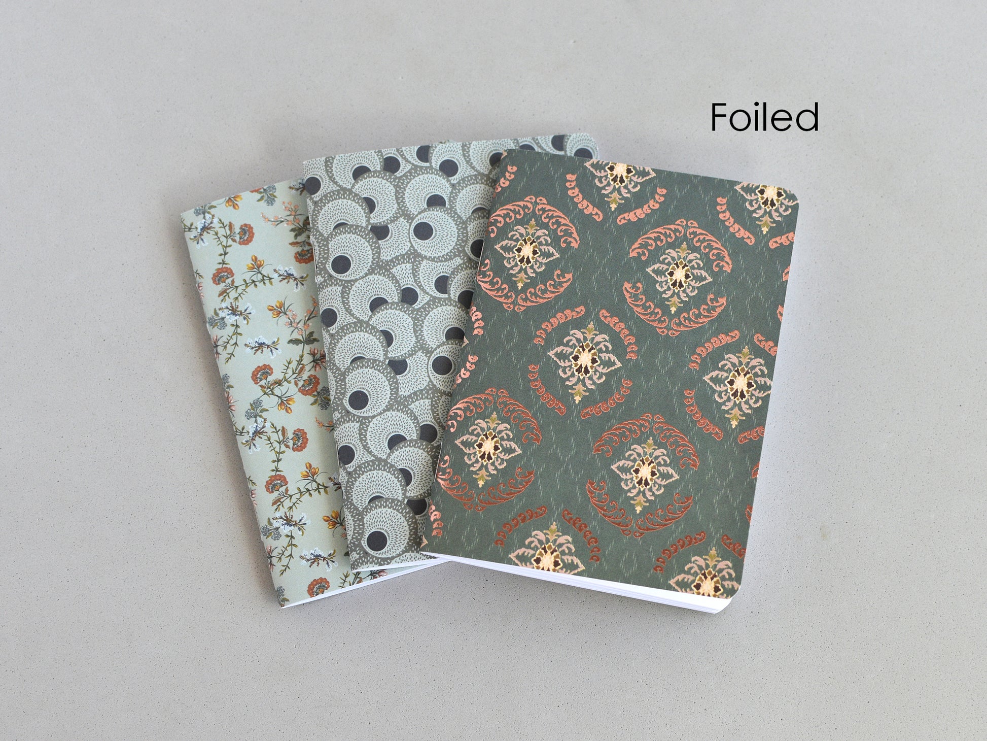 Three small patterned notebooks or journals fanned out on a light gray background, with the front cover showing a metallic foil floral and geometric design and the word 'Foiled' printed in the upper right.