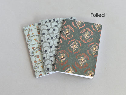 Three small patterned notebooks or journals fanned out on a light gray background, with the front cover showing a metallic foil floral and geometric design and the word 'Foiled' printed in the upper right.