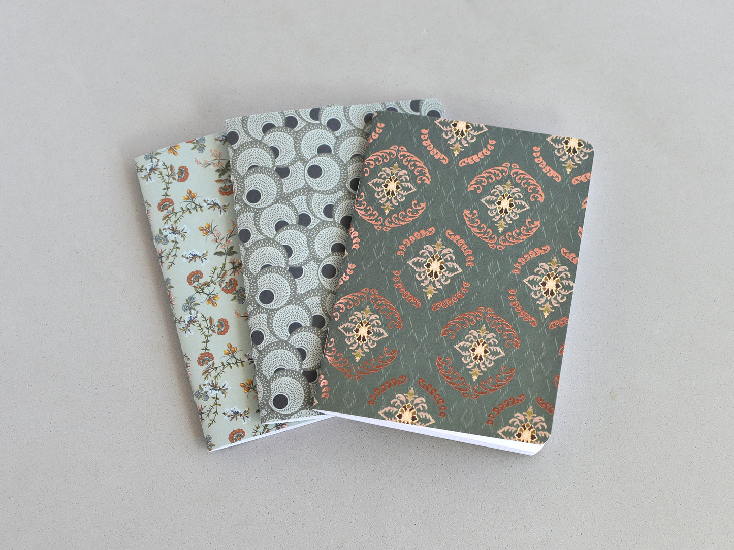 Three small notebooks or journals fanned out on a light gray background. The notebook on top has a dark green damask pattern with gold and rust accents, the middle notebook has a gray and black circular pattern, and the bottom notebook has a pale green floral pattern.