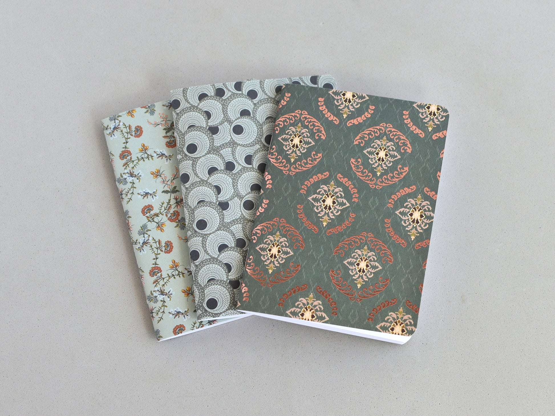 Three small notebooks or journals fanned out on a light gray background. The notebook on top has a dark green damask pattern with gold and rust accents, the middle notebook has a gray and black circular pattern, and the bottom notebook has a pale green floral pattern.